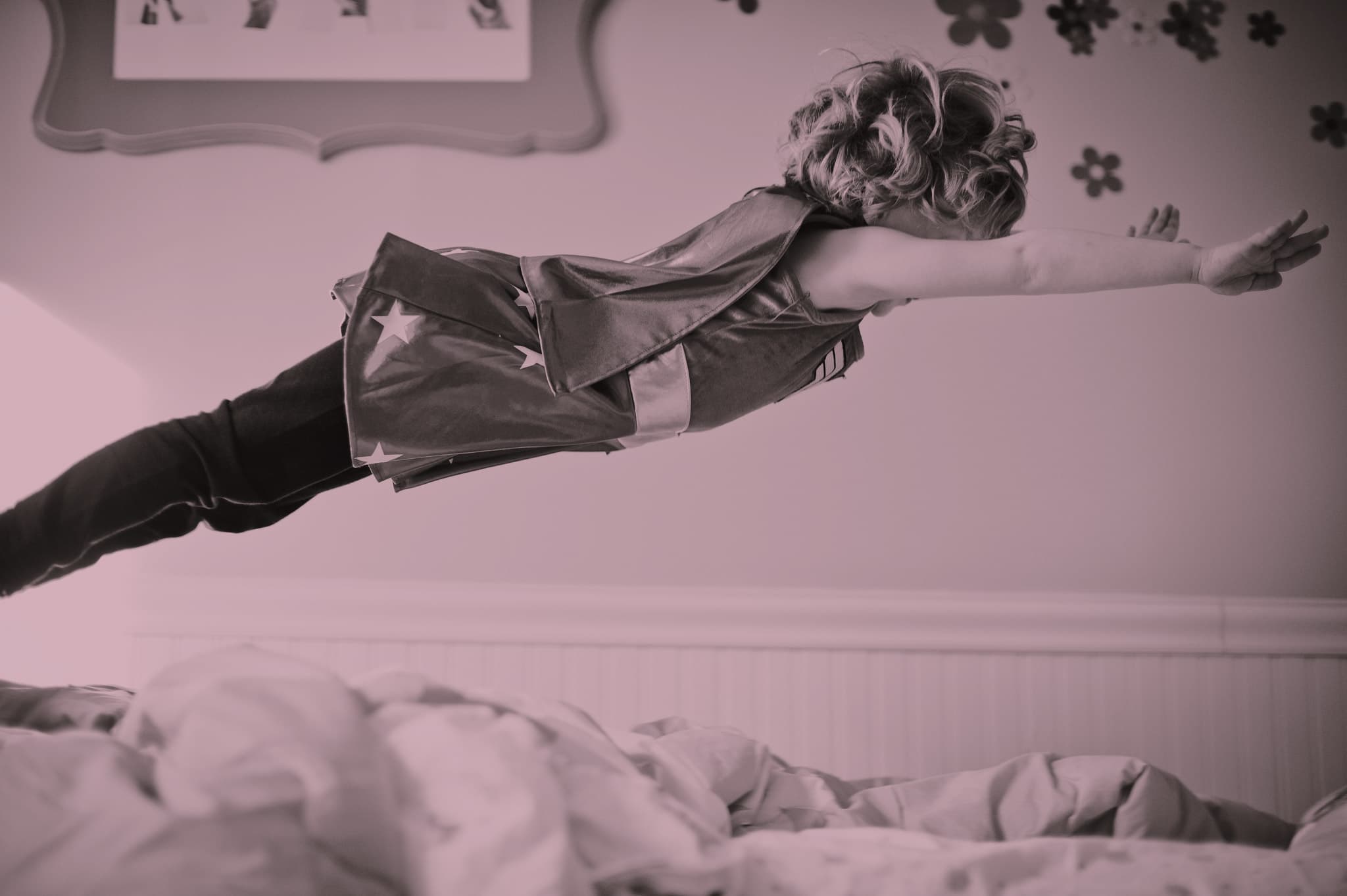 A child with curly hair, wearing a superhero cape, is captured mid-air as they leap from a bed, arms outstretched in a flying pose. The room is decorated with flower stickers on the wall.