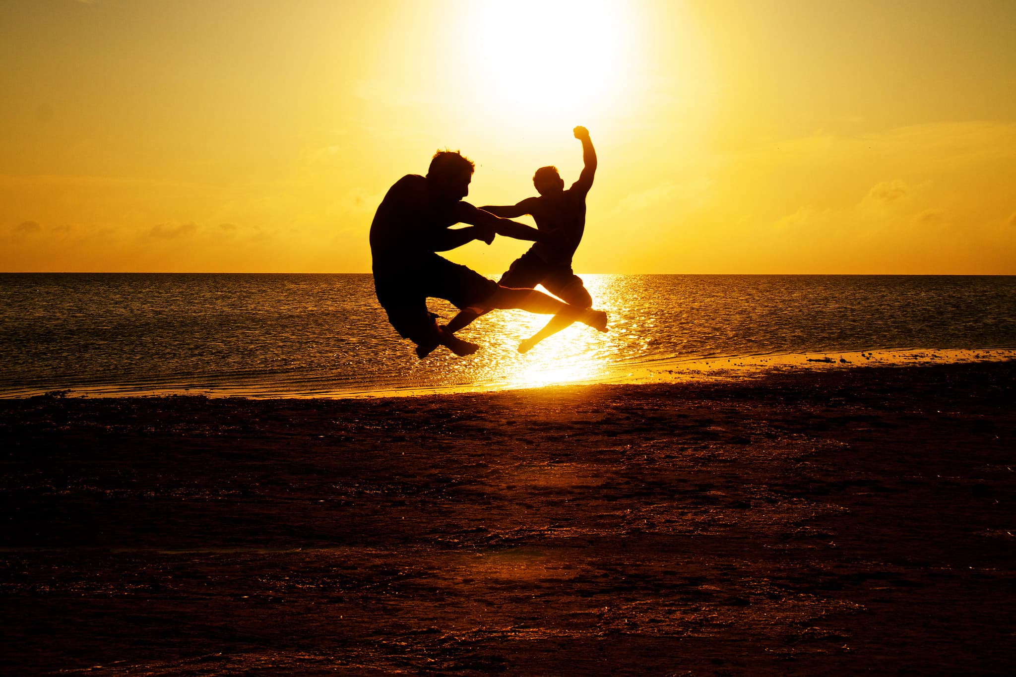 Two silhouetted figures are captured mid-air in dynamic poses against a golden sunset backdrop on a beach. The ocean glistens with the suns reflection, creating a dramatic and vibrant scene.