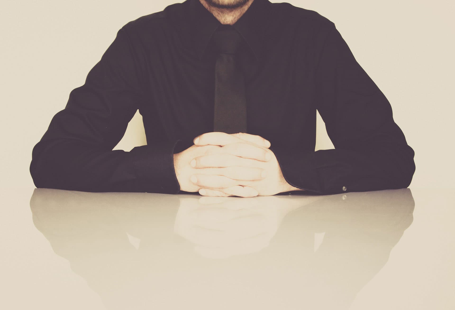 A person in a black shirt and tie sits at a reflective table with hands clasped together, against a neutral background.