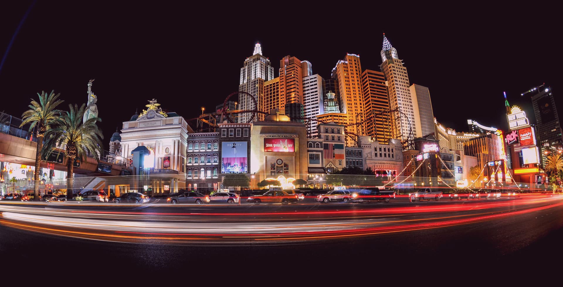 Night view of a brightly lit cityscape featuring tall buildings styled like New York City landmarks. A blend of neon lights and blurred car lights create dynamic streaks in the foreground, adding a sense of motion.