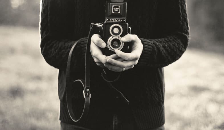 A person in a knitted sweater holding a vintage Argoflex camera with both hands. The background is softly blurred, suggesting an outdoor setting. The image is in black and white, capturing a nostalgic feel.
