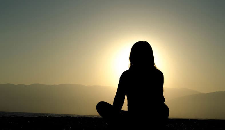 Silhouette of a person meditating by the water during sunset. The sun is low on the horizon, casting a warm glow. Mountains are visible in the background, and the persons figure is centered, sitting cross-legged.