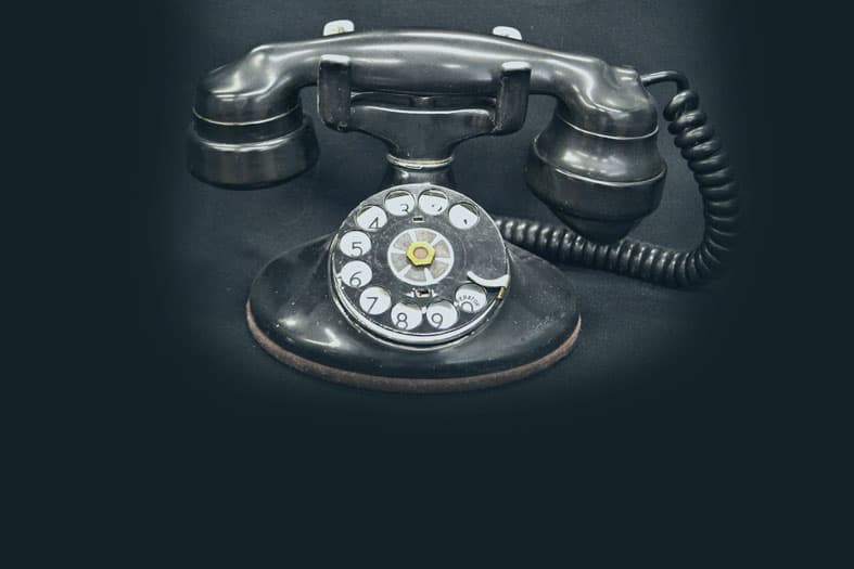 A vintage black rotary dial telephone with a classic handset and coiled cord sits against a dark background. The numbers 0 to 9 encircle the dial in white.