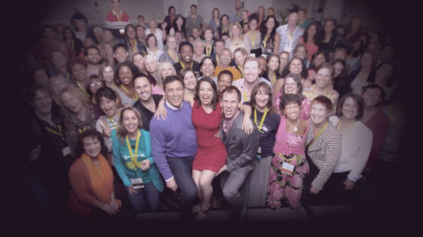 A large group of diverse individuals gathered closely, smiling and posing for the camera in a joyful setting. A few people in the front center are sitting on a platform, with many wearing conference lanyards.