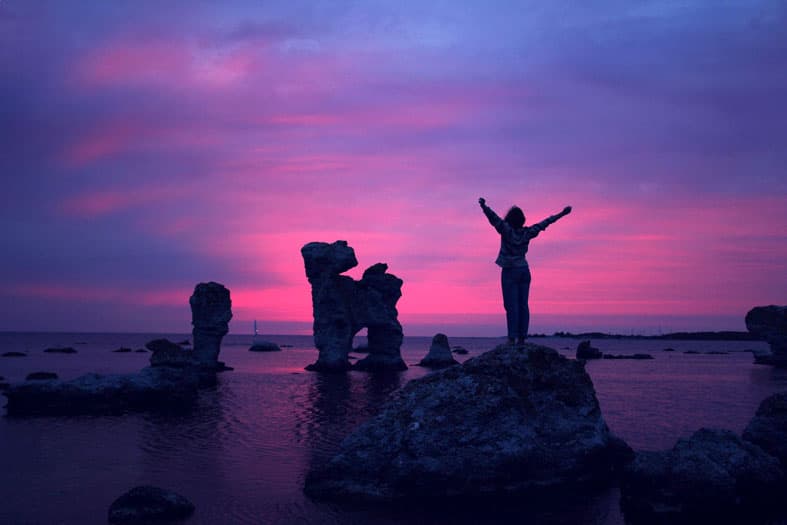 A person stands triumphantly on a rock by the sea, silhouetted against a vivid pink and purple sunset sky. Several large rock formations protrude from the water in the background.