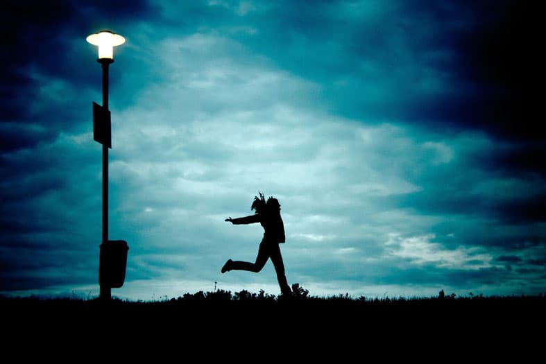 A silhouette of a person joyfully leaping in the air under a streetlight, with a dramatic cloud-covered sky in the background.