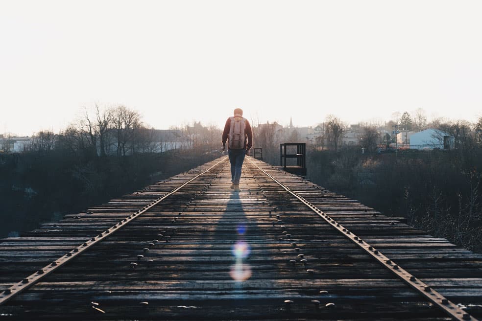 A person with a backpack walks alone on a deserted railway bridge, surrounded by trees and distant buildings, under a clear sky with the sun creating a lens flare on the wooden planks.