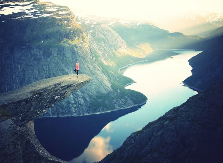 A person stands on the edge of a rocky cliff, with a vast view of a serene lake below and surrounding mountains under a hazy sky. The scene conveys a sense of adventure and tranquility in a breathtaking natural landscape.