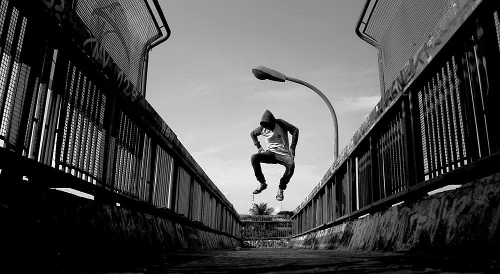 A person wearing a hoodie and jeans performs a jump on a pedestrian bridge. The silhouette contrasts against the bright sky, with metal railings on either side and a streetlamp in the background. Black and white image.