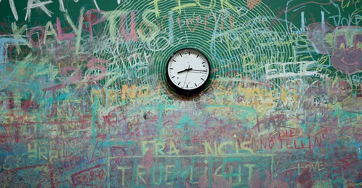 A wall clock is centered against a chalkboard covered in colorful chalk doodles, scribbles, and words. The artwork creates a vibrant, chaotic backdrop of overlapping colors and shapes.
