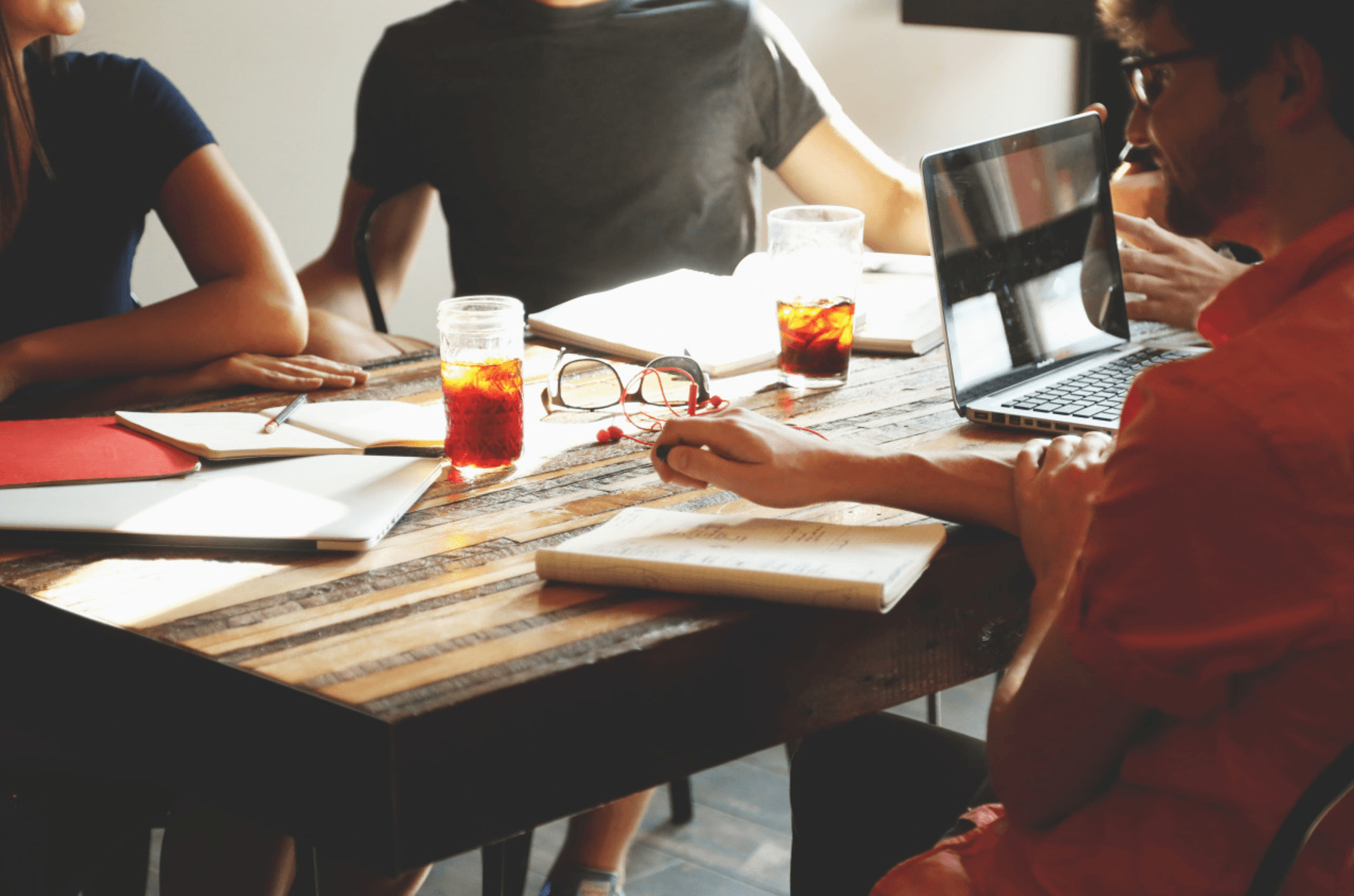 A group is sitting around a wooden table, having a meeting. There are notebooks, a laptop, and glasses of iced tea on the table. One person is holding a pen, and sunlight is streaming in, creating a bright and informal atmosphere.