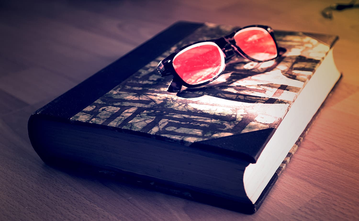 A closed, vintage-looking book with a patterned cover lies on a wooden surface. A pair of sunglasses with red-tinted lenses is placed on top of the book, casting a warm glow in the dim lighting.