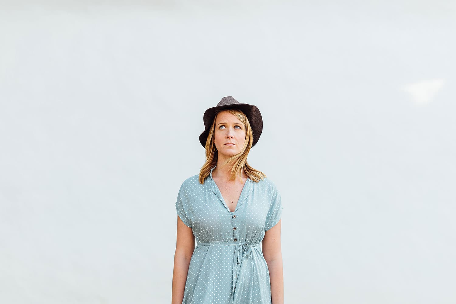 A woman stands against a plain light background, wearing a blue polka dot dress and a dark hat. She looks off to one side with a neutral expression.