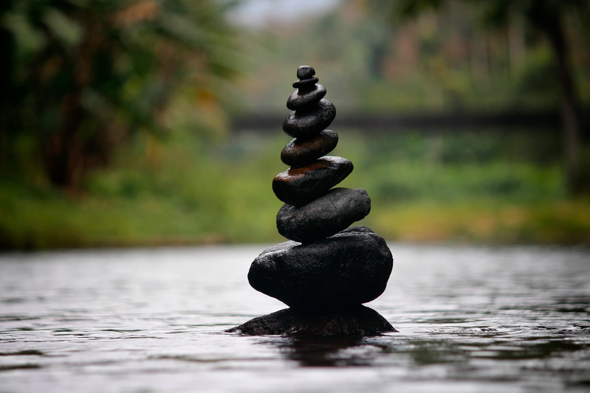 A stack of smooth stones balanced on each other in a calm, shallow stream. The background is blurred with green foliage, creating a peaceful, natural atmosphere.