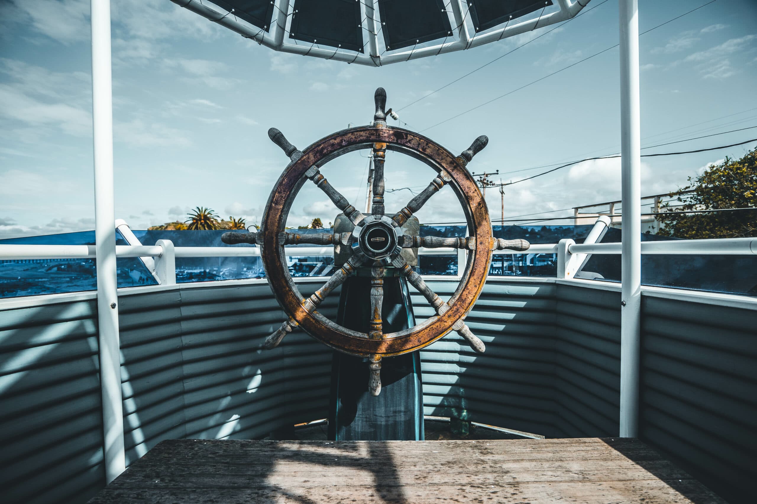 A wooden ships wheel on a raised deck with a nautical theme. The sky is partly cloudy, and there are trees visible in the background. The scene suggests a maritime setting.
