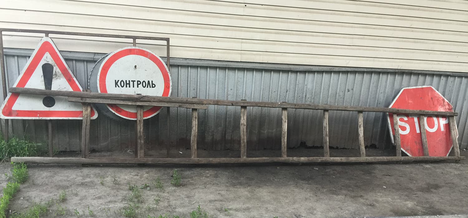 A wooden ladder rests against a corrugated metal wall. Beside it are three road signs: a triangular sign with an exclamation mark, a circular sign with text, and a red octagonal sign with STOP written on it.