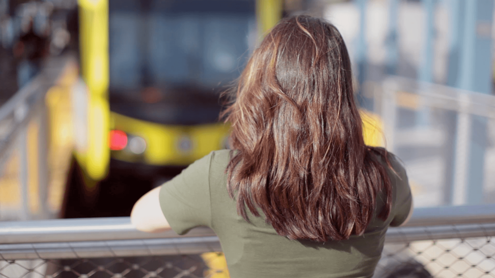 A person with long brown hair, wearing a green shirt, stands leaning on a railing and looks at an approaching yellow tram at a station. The background is slightly blurred.
