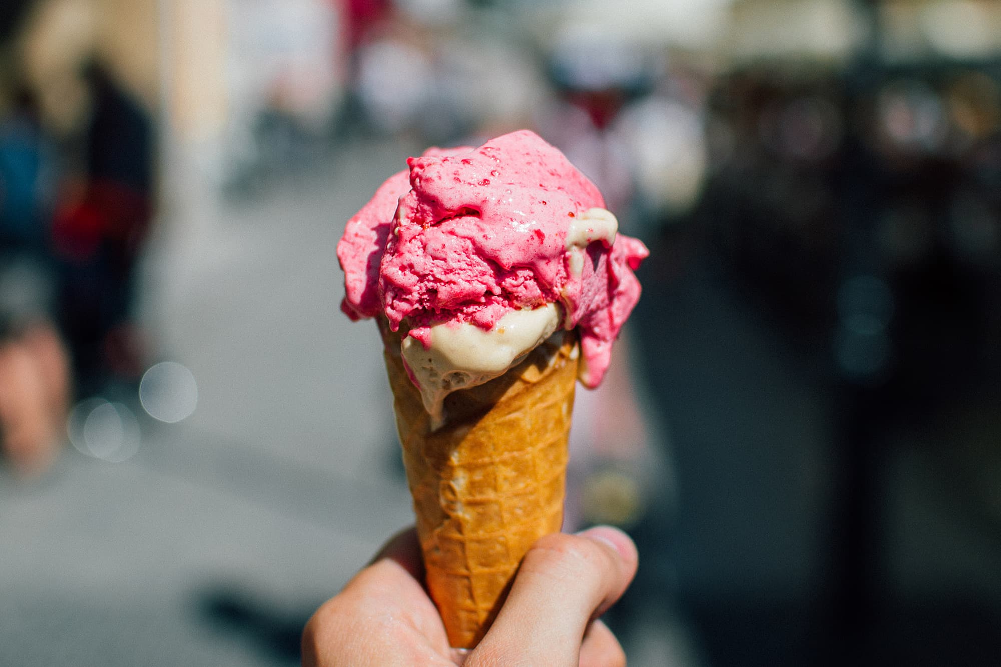 A hand holding an ice cream cone with two scoops: one bright pink and one cream-colored. The background is blurred, suggesting an outdoor setting on a sunny day.
