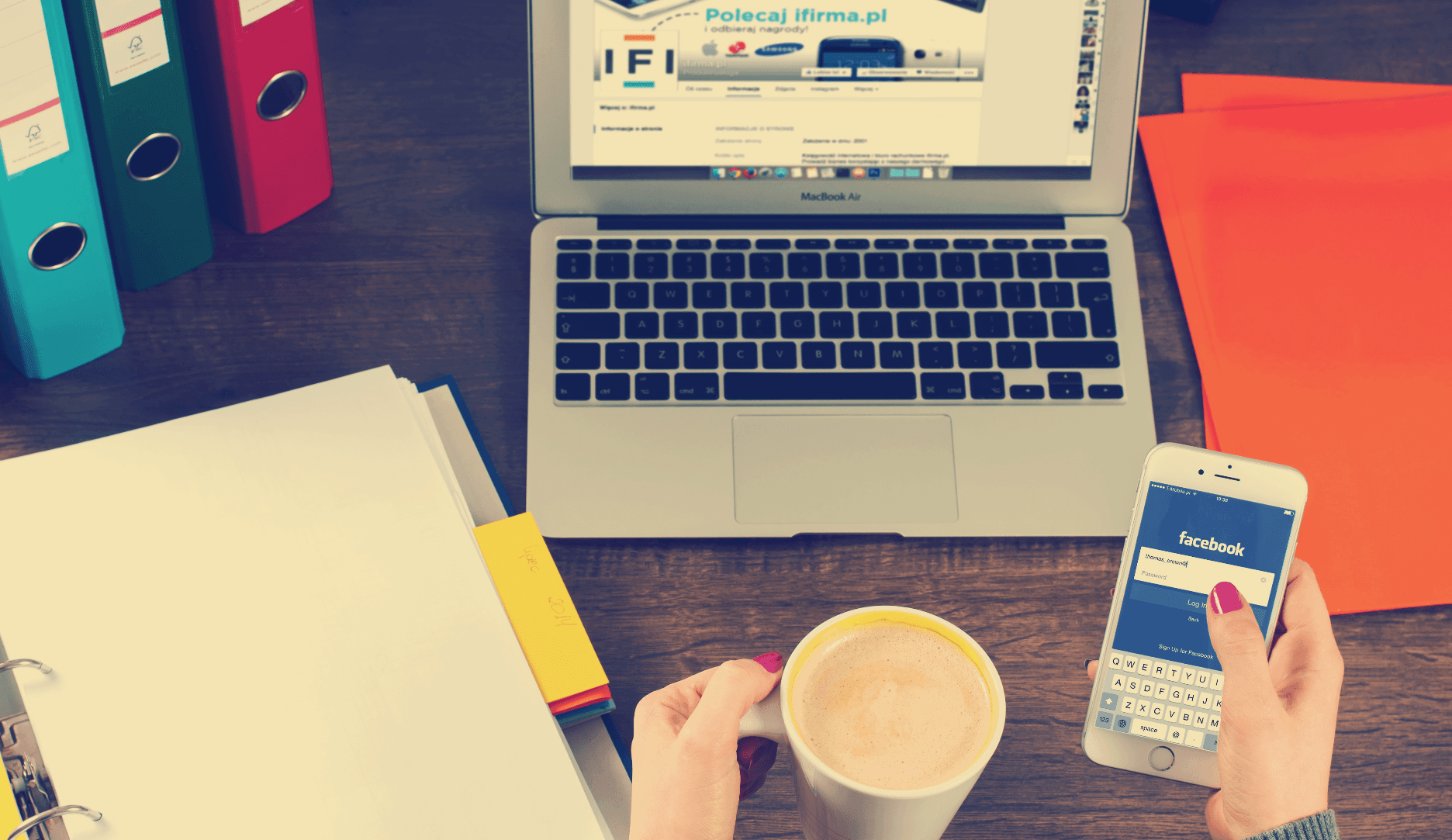 A person holds a cup of coffee in one hand and a smartphone in the other, displaying the Facebook login page. A laptop is open in front of them on a wooden desk, surrounded by colorful binders and documents.
