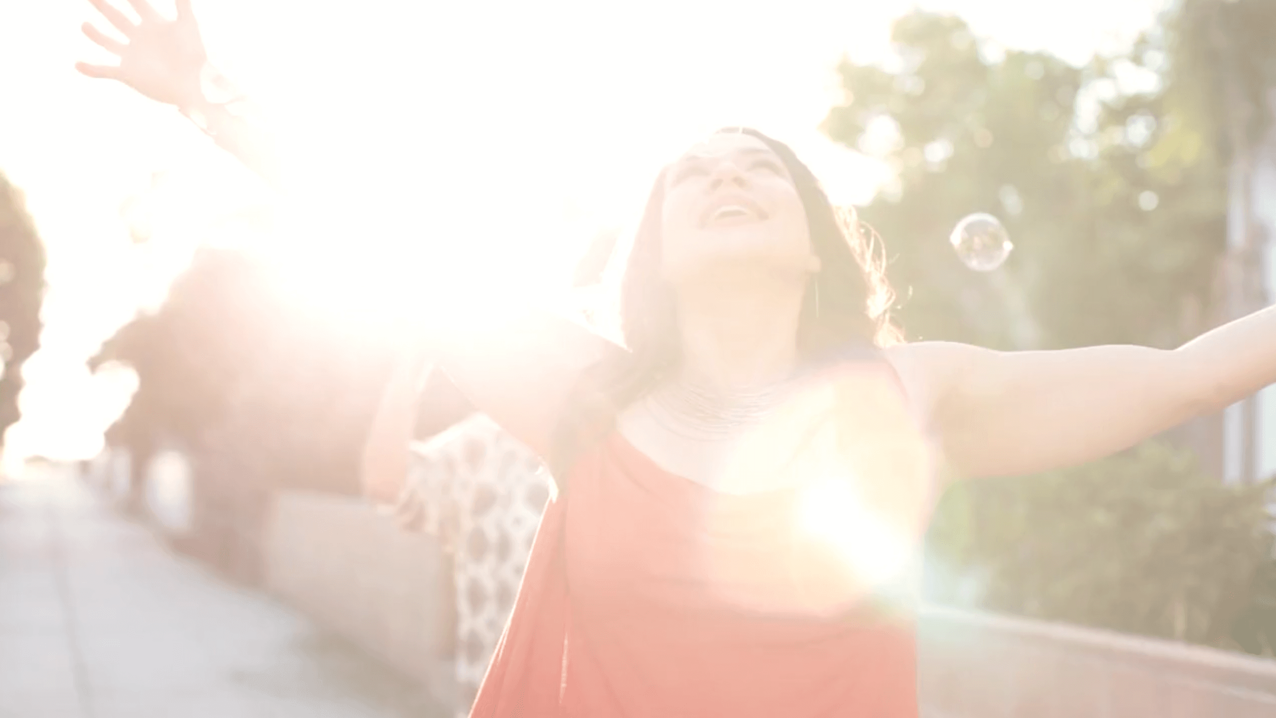A woman in a red dress stands with arms outstretched, basking in bright sunlight on a street with trees. A bubble floats nearby, and the photo has a soft, dreamy atmosphere.