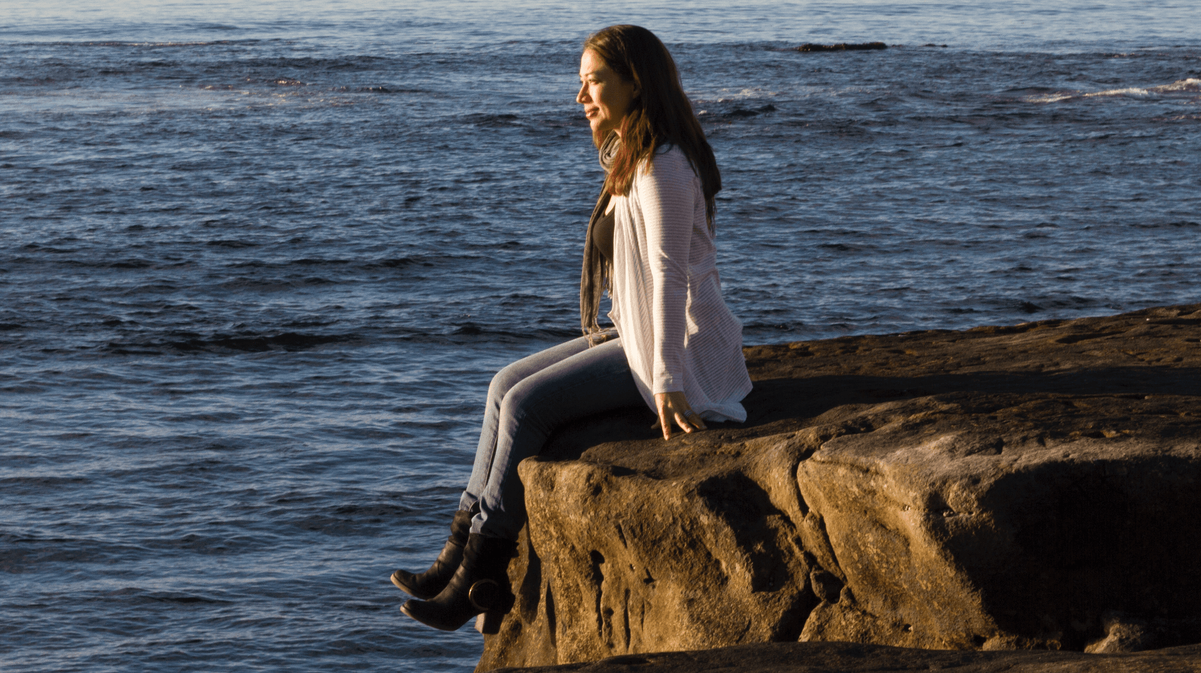 A person sits on a rocky ledge overlooking a calm ocean, wearing a white cardigan, blue jeans, and black boots. The sun casts a warm glow on their face, creating a peaceful and serene scene.