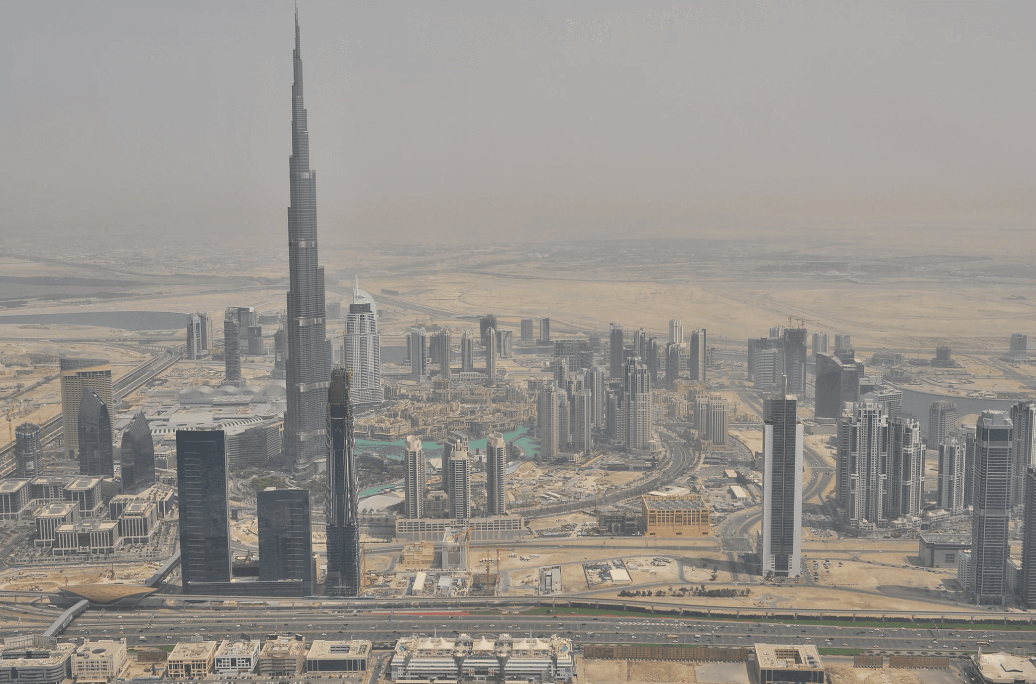 Aerial view of Dubais skyline featuring the Burj Khalifa surrounded by modern skyscrapers and desert landscape. The cityscape includes various tall buildings and urban structures with clear skies above.