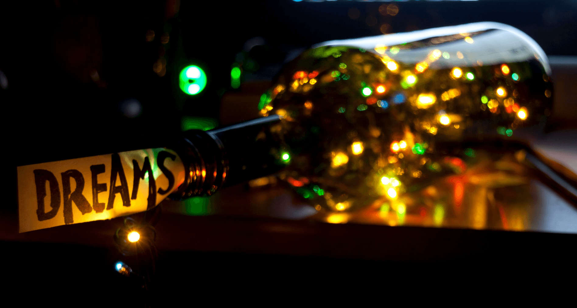 A glass bottle filled with colorful string lights lies on its side in a dimly lit room. A paper tag reading DREAMS is attached to the bottles neck, with lights twinkling in the background.