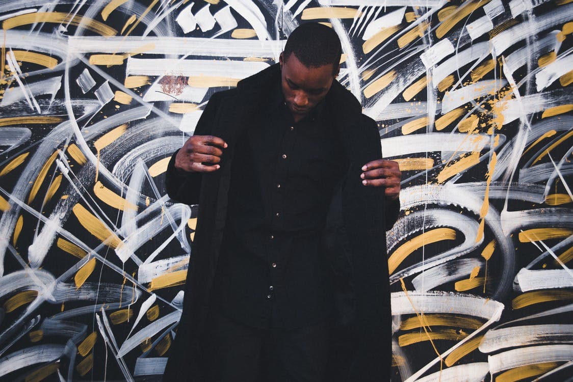 A person in a black outfit stands in front of a dynamic black, white, and yellow abstract mural. They are adjusting their jacket, looking down slightly, creating a striking contrast with the vivid background.