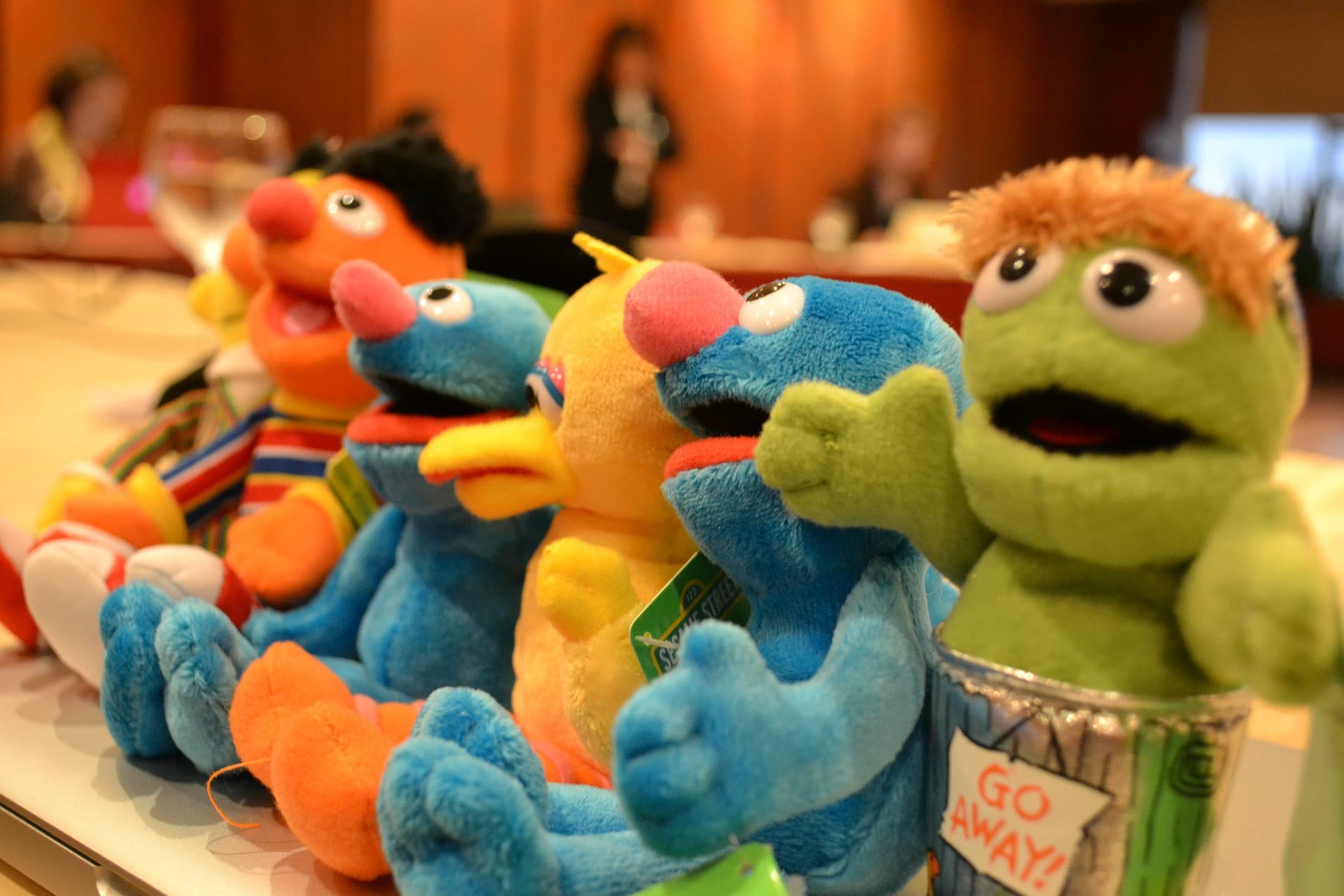A row of colorful plush toys resembling popular television characters is lined up on a table. The toys include a yellow beaked character, a green figure in a trash can marked Go Away!, and others with distinctive features.