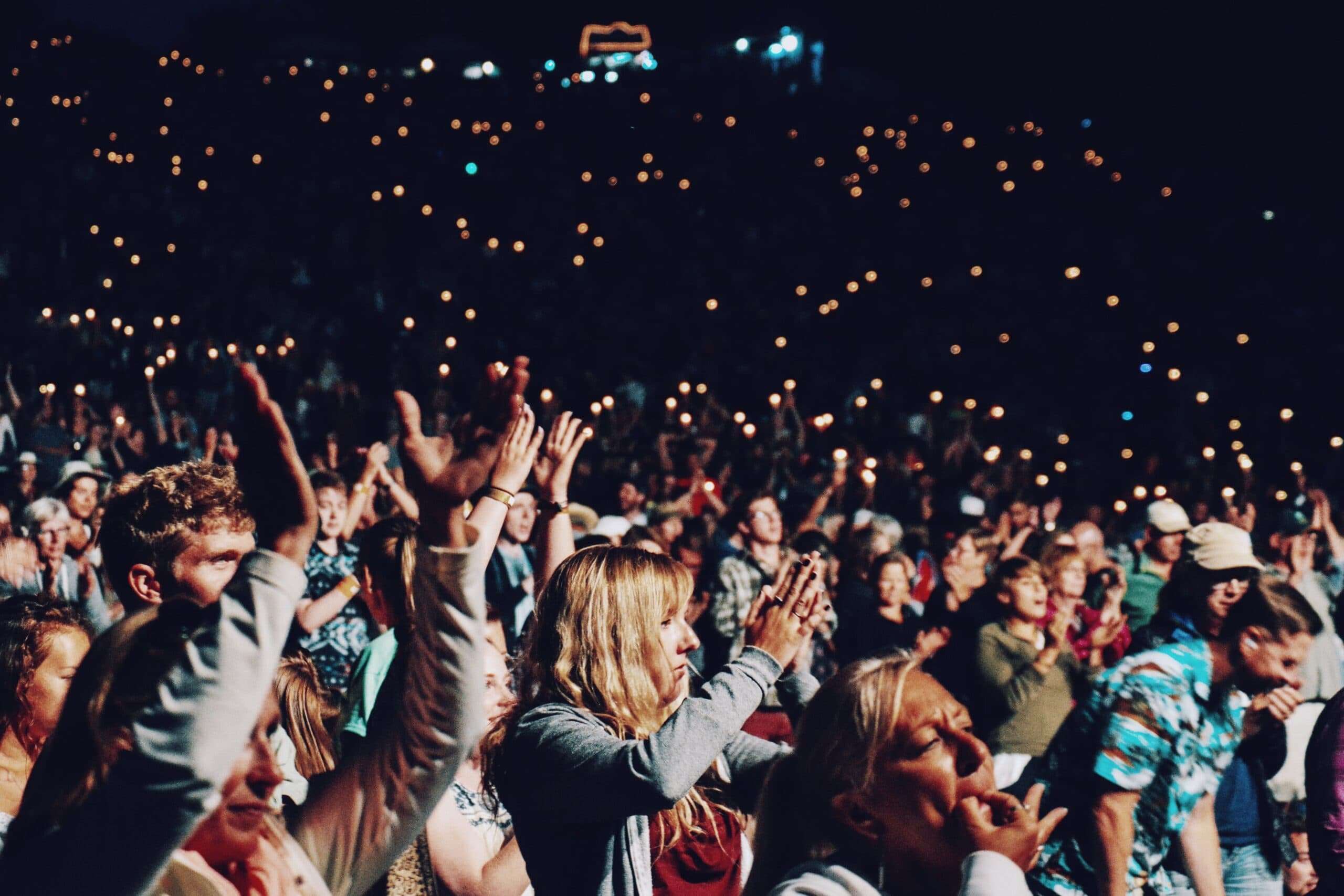 A large crowd at a nighttime concert, clapping and holding lit phones or lighters, creating a sea of tiny lights. The audience is diverse, with people standing closely together, adding to the lively atmosphere.