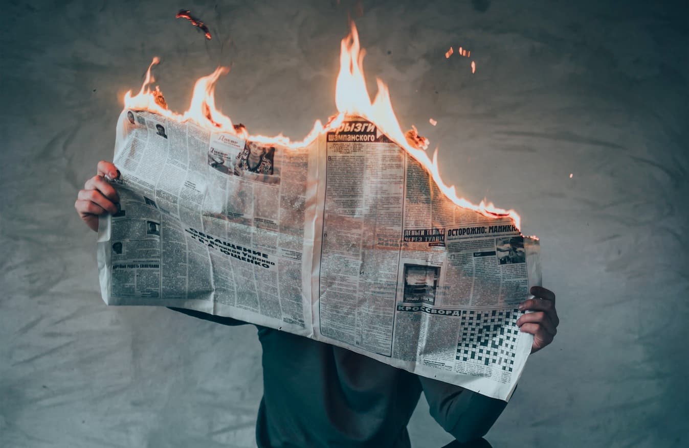 A person holds an open newspaper engulfed in flames, with text in a foreign language visible. The background is a plain gray wall, and the persons face is obscured by the newspaper.