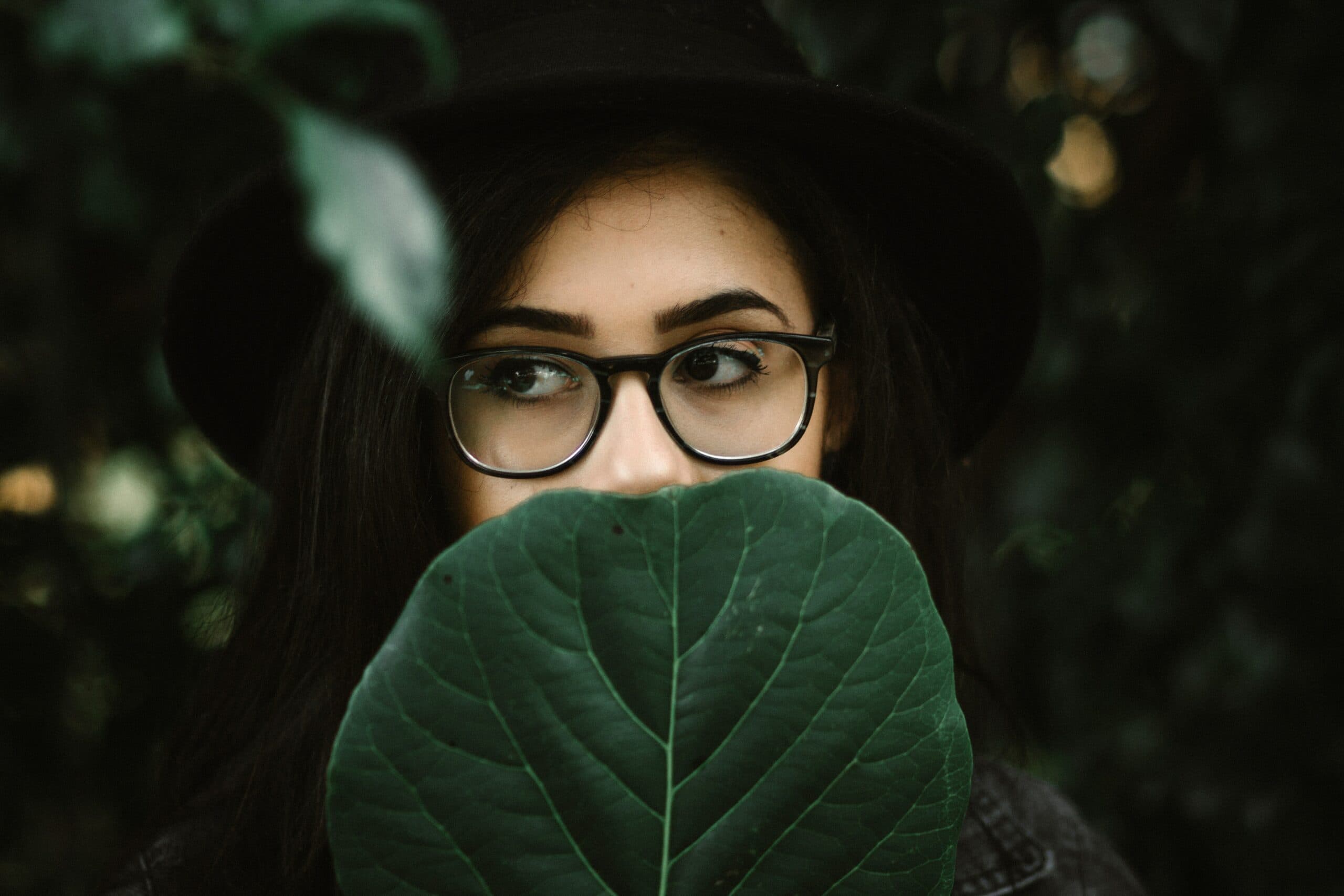 A person with long dark hair, wearing glasses and a black hat, is partially obscured by a large green leaf held in front of their face. The background is filled with greenery.
