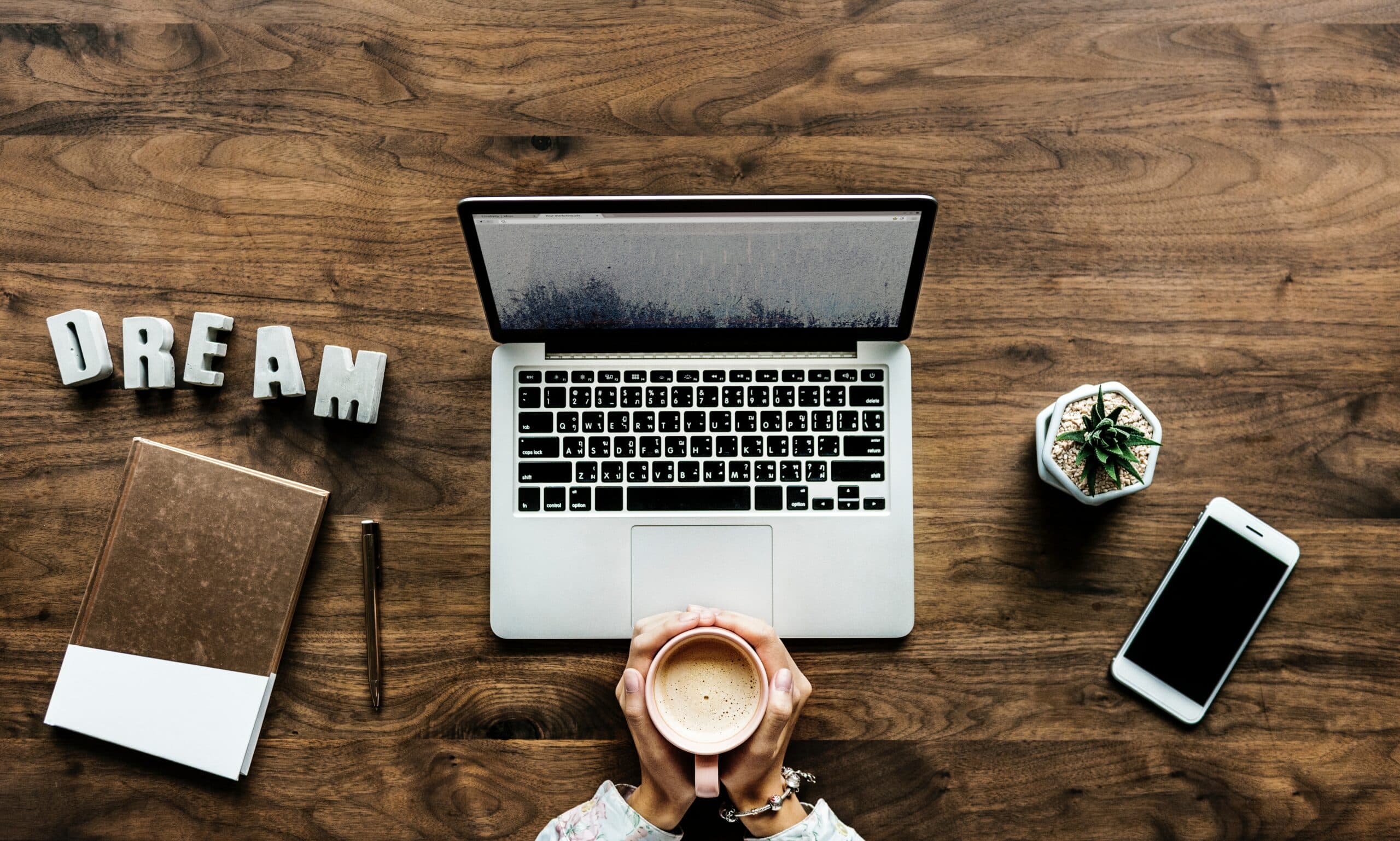 A person holds a cup of coffee at a wooden desk featuring a laptop, a notebook with a pen, a small potted plant, a smartphone, and decorative letters spelling DREAM.