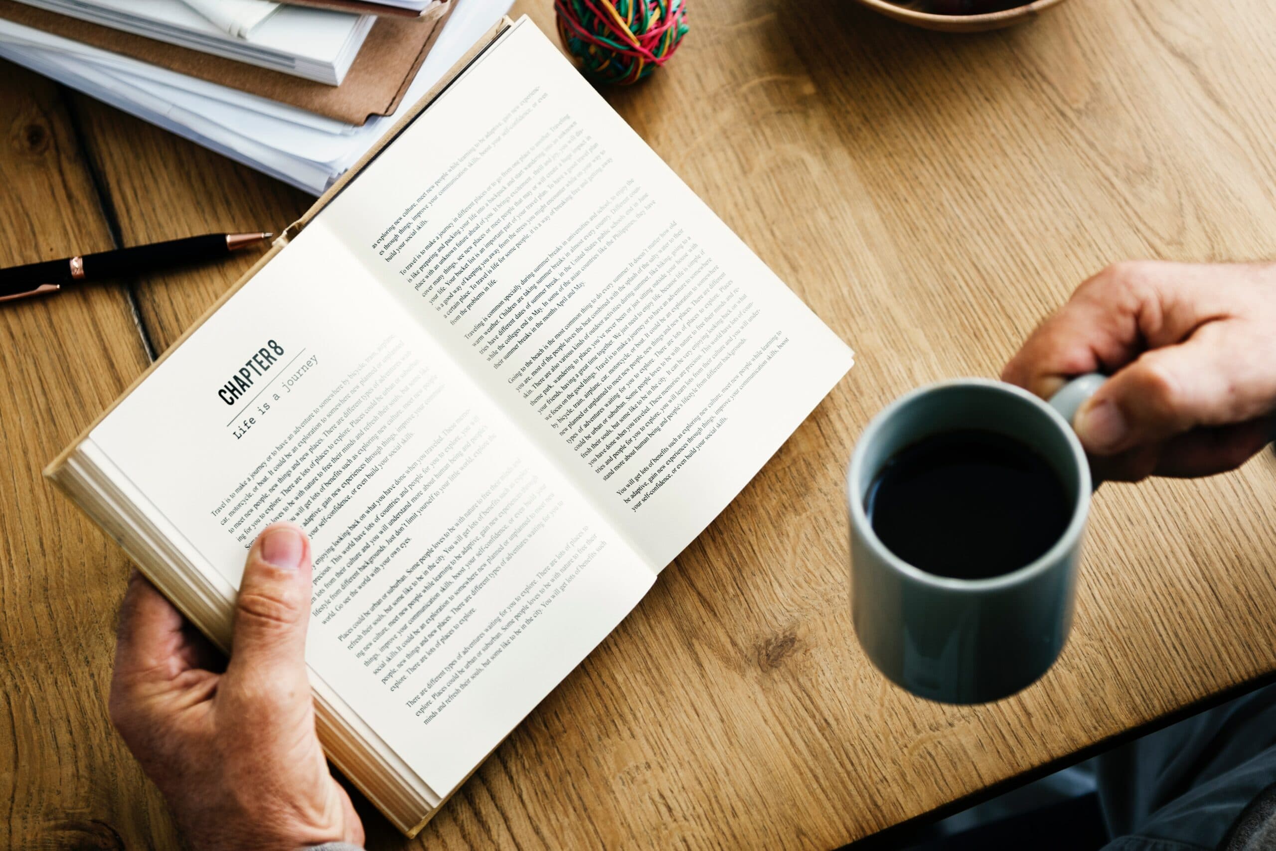 A person is holding a book open to a chapter titled Life and Mystery while holding a mug of coffee in the other hand. The scene is set on a wooden table with some pens and papers in the background.