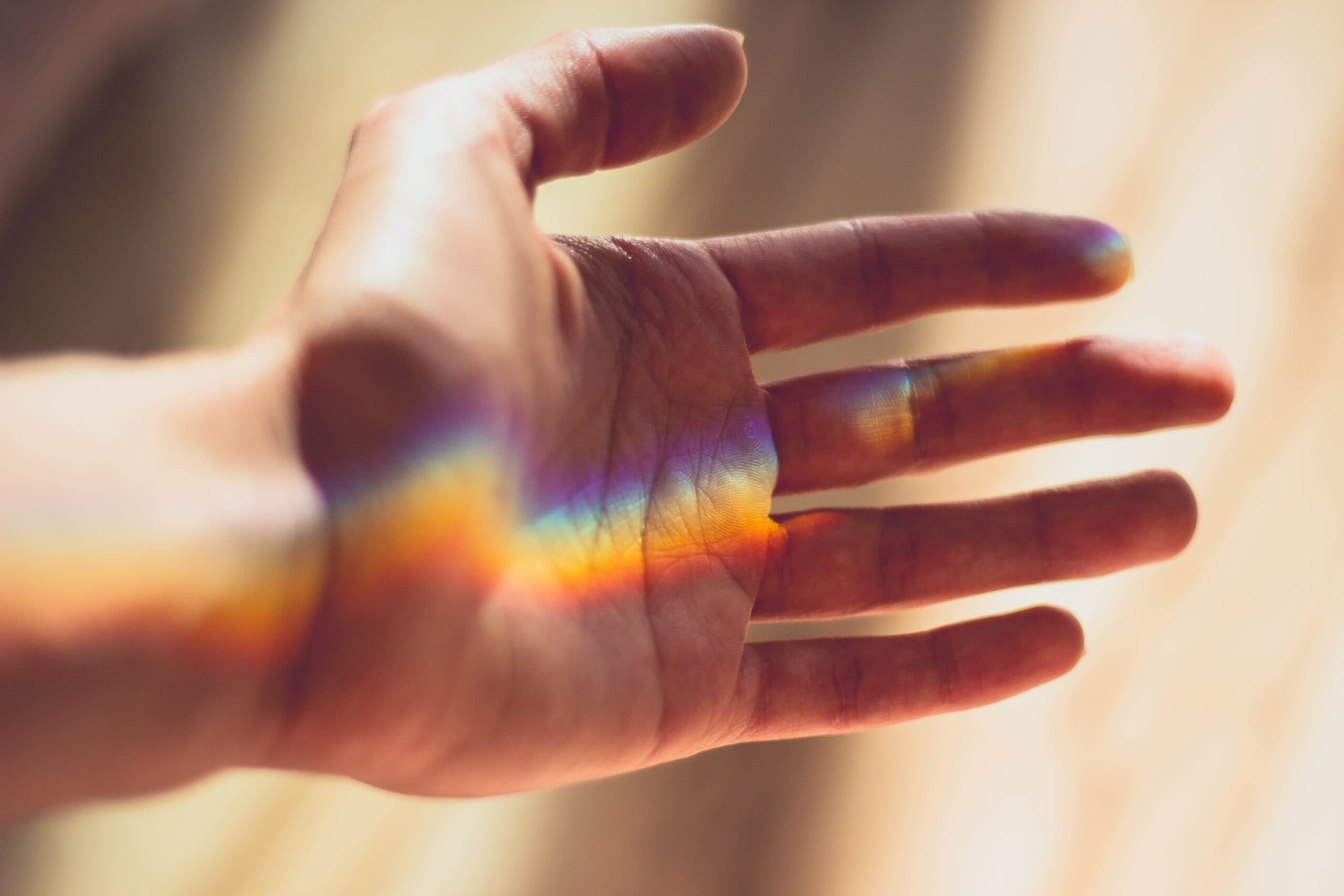 A hand is outstretched with sunlight creating a rainbow across the palm. The background is softly blurred in warm tones, enhancing the effect of the colorful spectrum on the skin.
