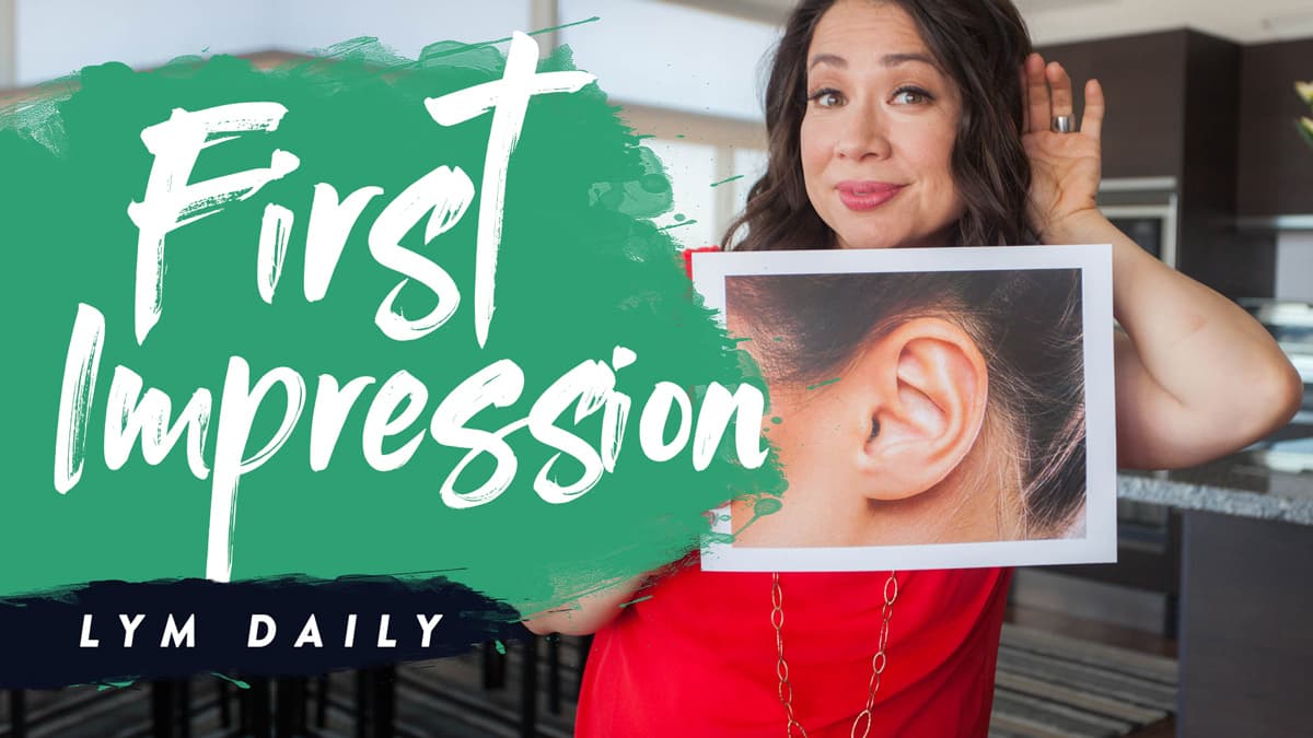 A woman in a red dress holds a picture of an ear next to her ear, with a playful expression. A green and white overlay reads First Impression LYM Daily.
