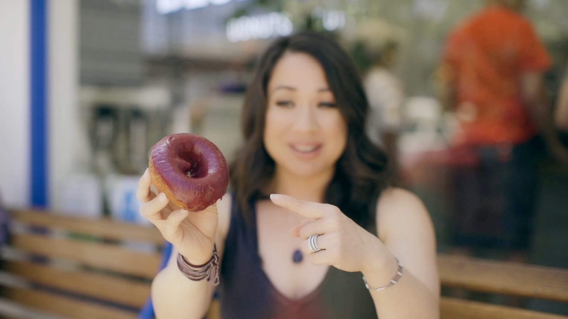 A person sitting on a bench holds a purple-glazed donut with a smile, pointing at it. The background is softly blurred, suggesting an outdoor setting.