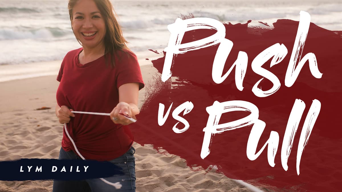 A woman in a red shirt smiles while standing on a beach, pulling on a rope. The words Push vs Pull are written in large, stylized white font on a dark red background. LYM Daily appears in the lower left corner.