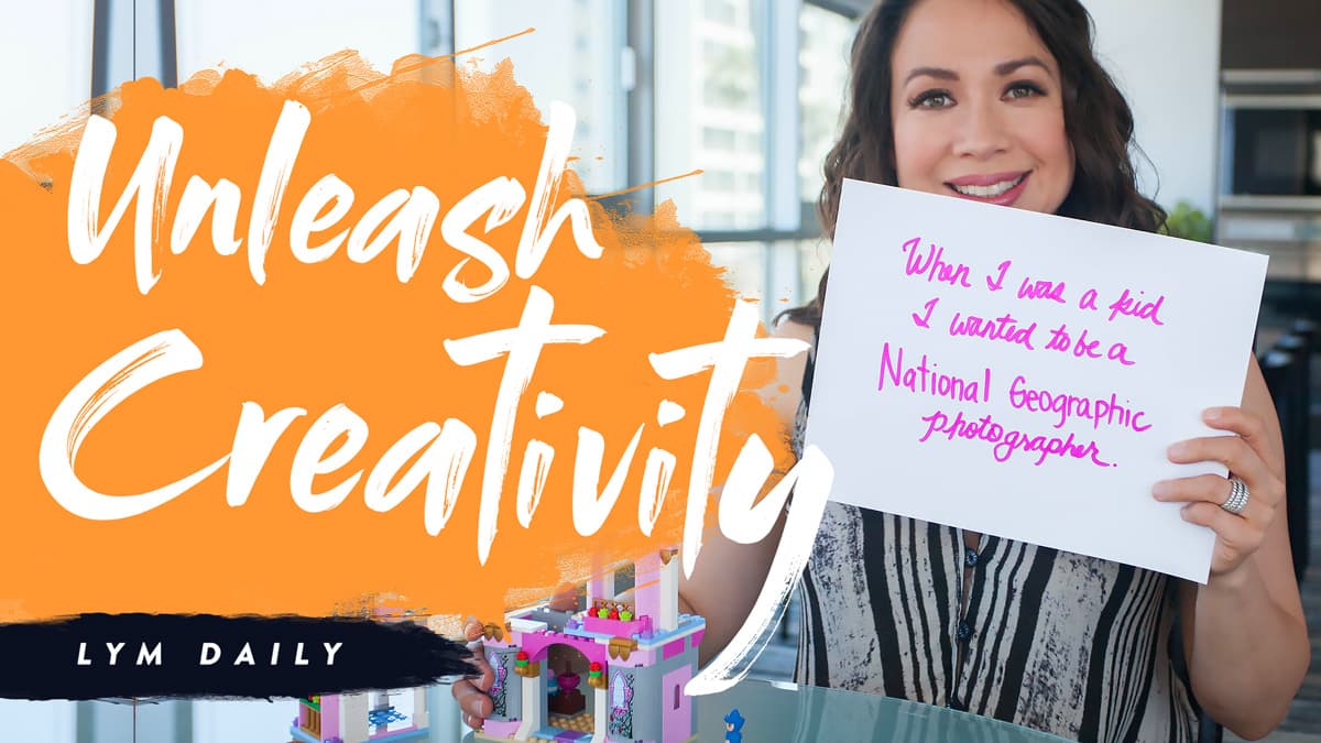 A woman in a brightly lit room holds a sign that reads, When I was a kid I wanted to be a National Geographic photographer. The text Unleash Creativity and LYM Daily are displayed in bold on the left side. Some colorful blocks are visible in the foreground.