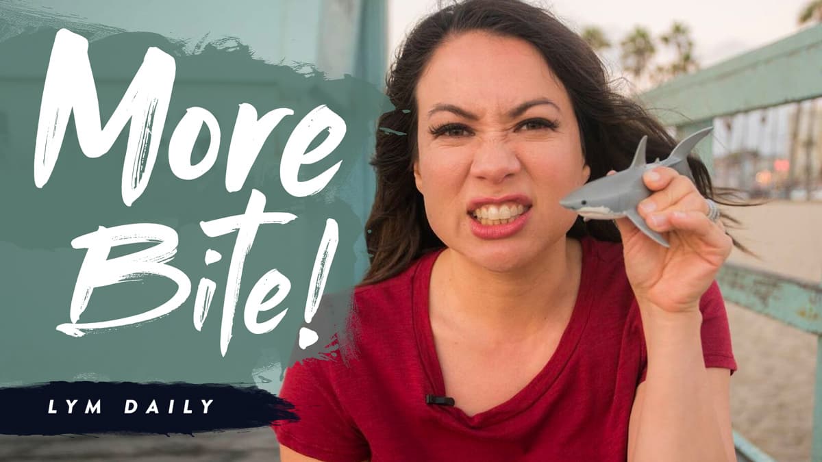 A woman in a red shirt holds a small toy shark near her face, making a playful grimace. The text More Bite! appears on the left, with LYM Daily below. The background shows a beach setting with palm trees.