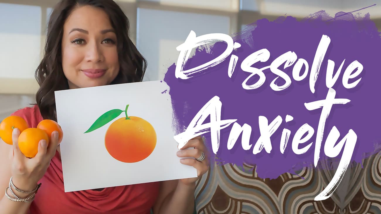 A woman holds three oranges in one hand and a picture of an orange in the other. The purple background features the text Dissolve Anxiety. She is smiling in a brightly lit room with patterned furniture.