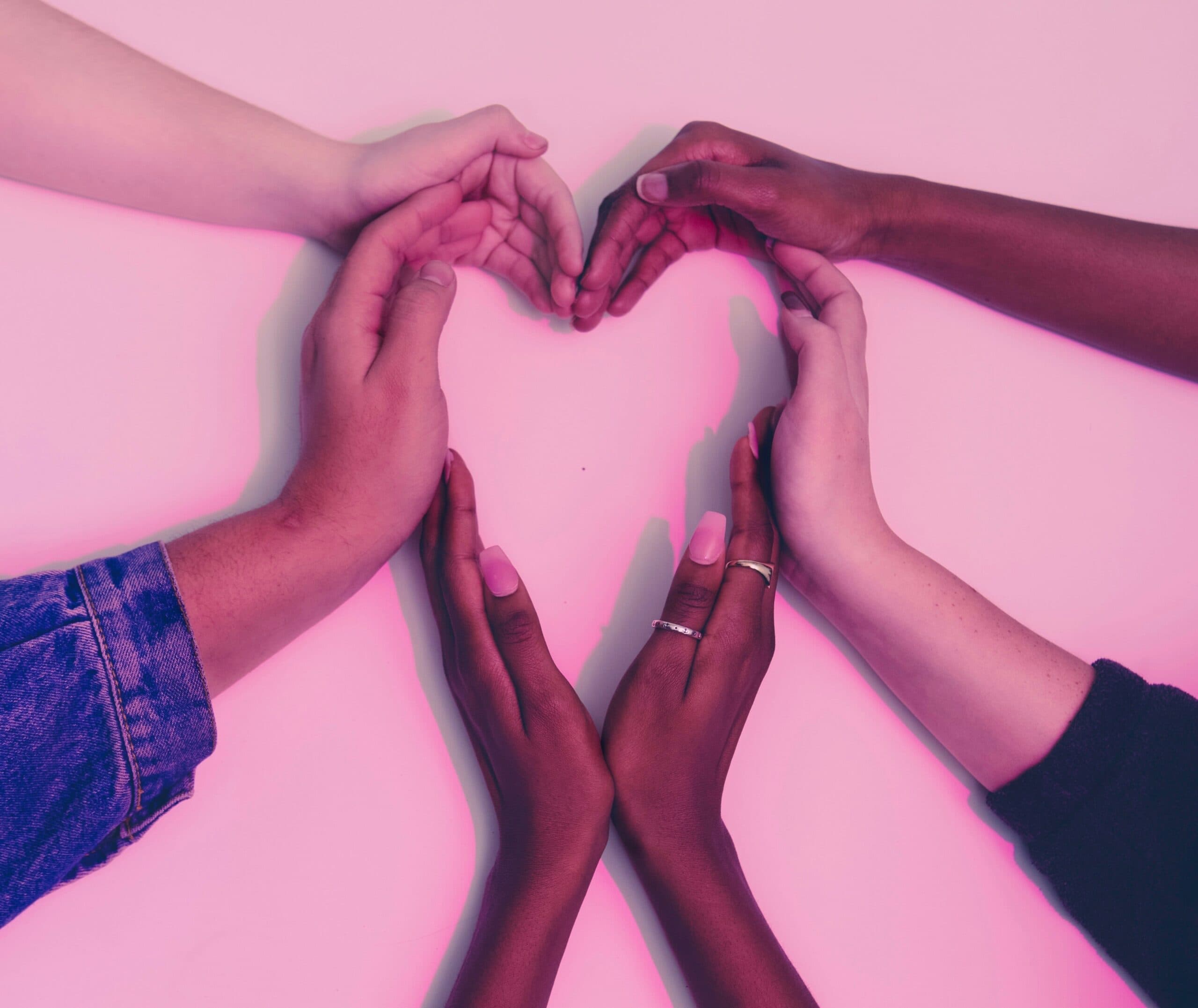 Six hands of diverse skin tones form the shape of a heart against a pink background. The hands are arranged symmetrically, with fingers touching to create the heart shape, symbolizing unity and love.
