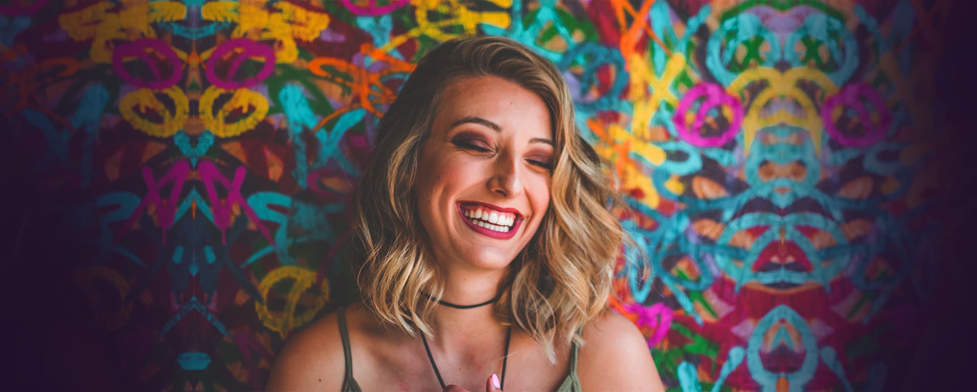 A woman with blonde hair, wearing a black choker and light-colored top, smiles brightly with closed eyes. She stands against a vibrant, colorful abstract mural, creating a joyful and lively atmosphere.