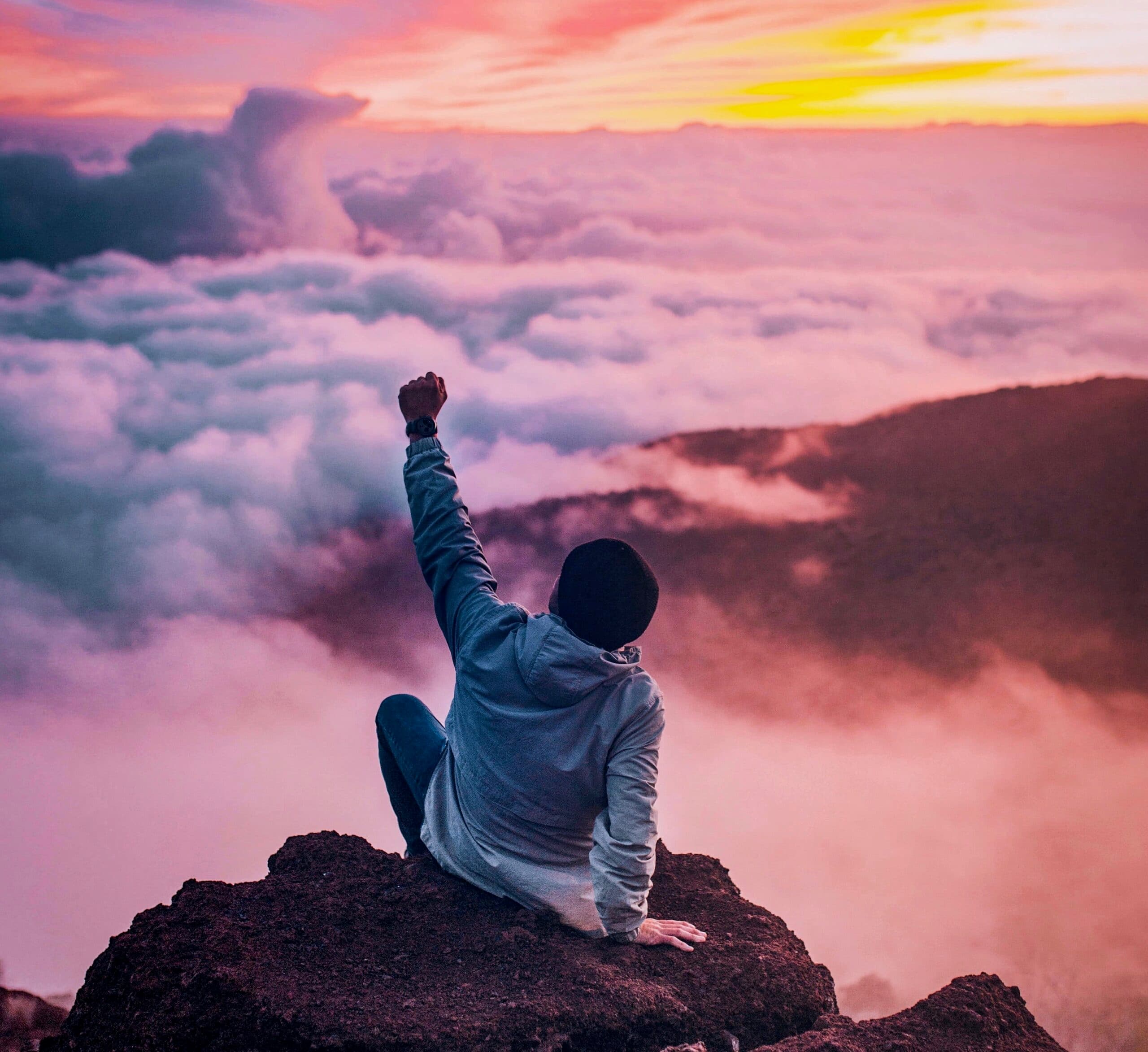 A person sits on a rocky peak above the clouds at sunset, raising a fist. The sky is filled with vibrant hues of pink, orange, and purple, creating a dramatic and inspiring scene.