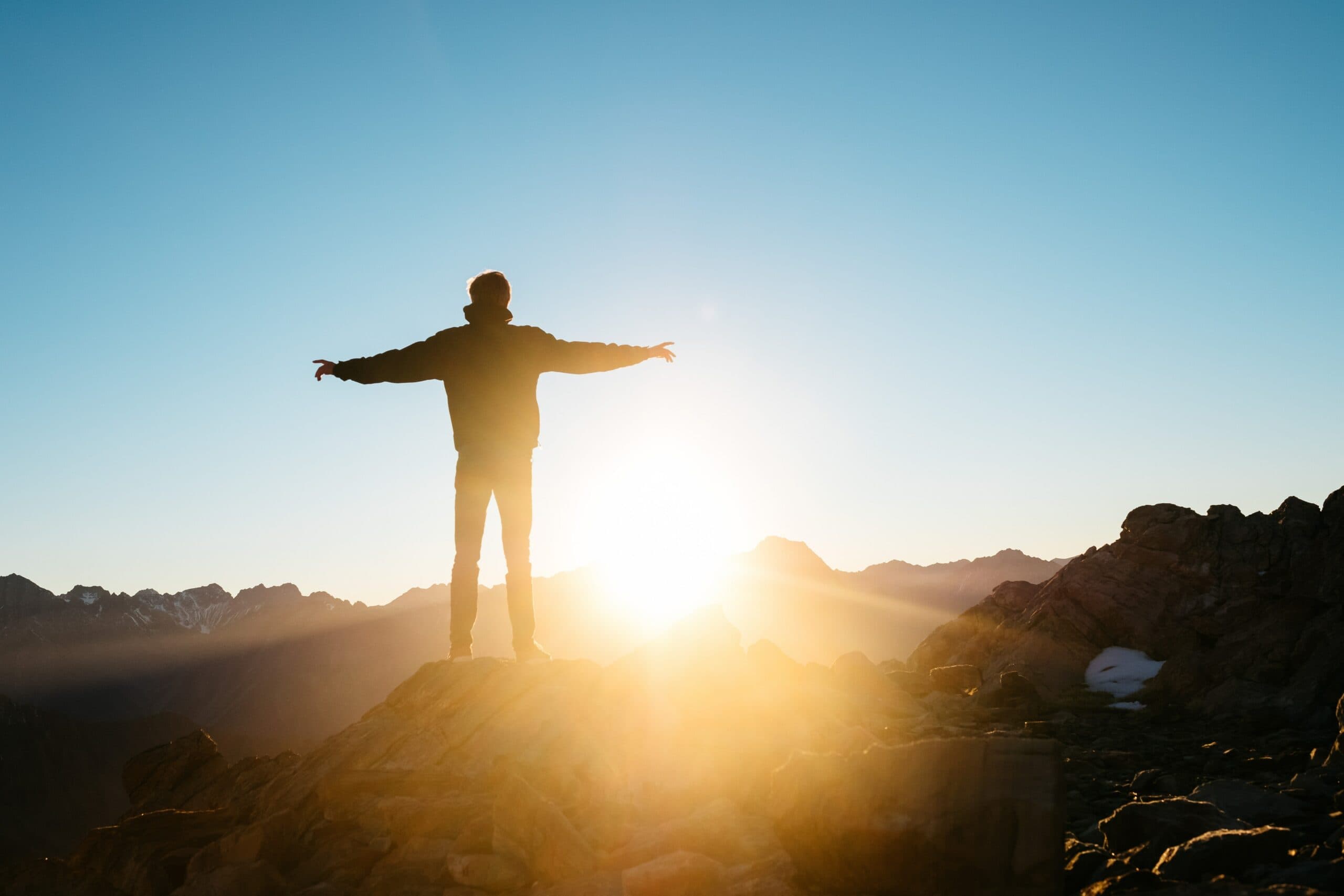 A person stands on a rocky mountaintop with arms outstretched, silhouetted against a bright sunrise. The horizon is framed by distant mountains under a clear blue sky.
