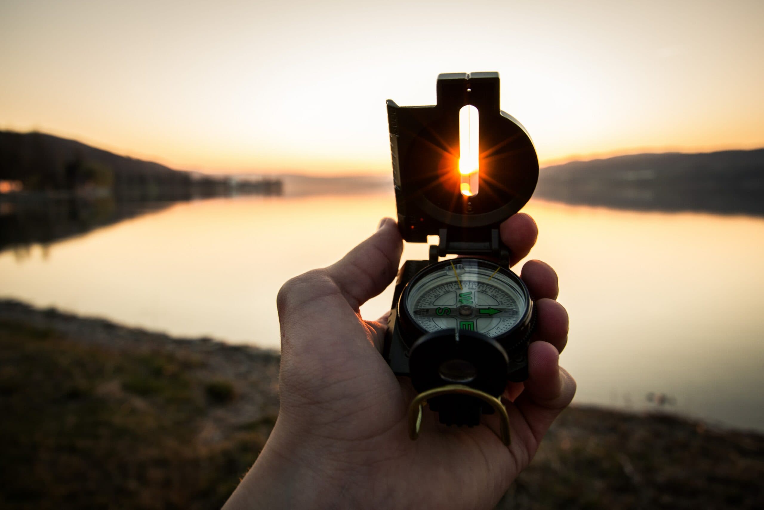 A hand holds a compass aligned with the setting sun, which shines through the compass sight. The background features a calm lake and silhouetted hills, bathed in golden light, creating a serene landscape.