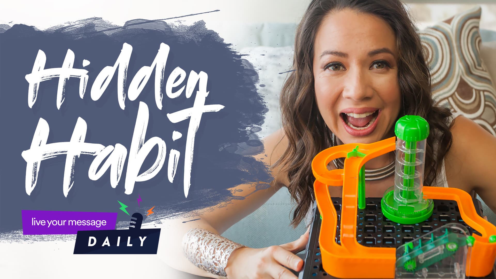 A person smiling enthusiastically holds an orange and green circuit-like contraption. Text on the image reads Hidden Habit with a logo live your message DAILY at the bottom left. A patterned cushion is visible in the background.