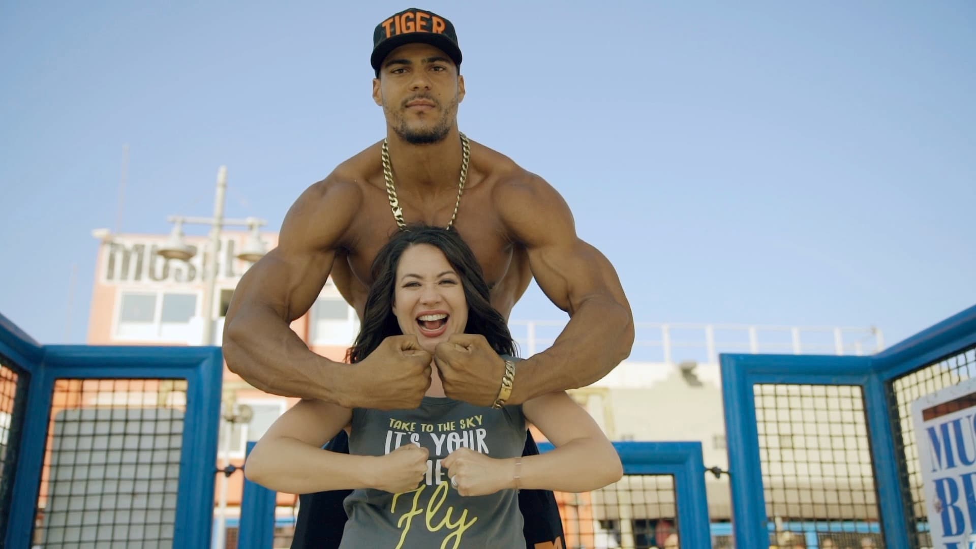 A muscular man wearing a cap and gold chain flexes his arms behind a smiling woman who is also flexing her arms. They are outdoors, with blue metal bars and a building in the background.