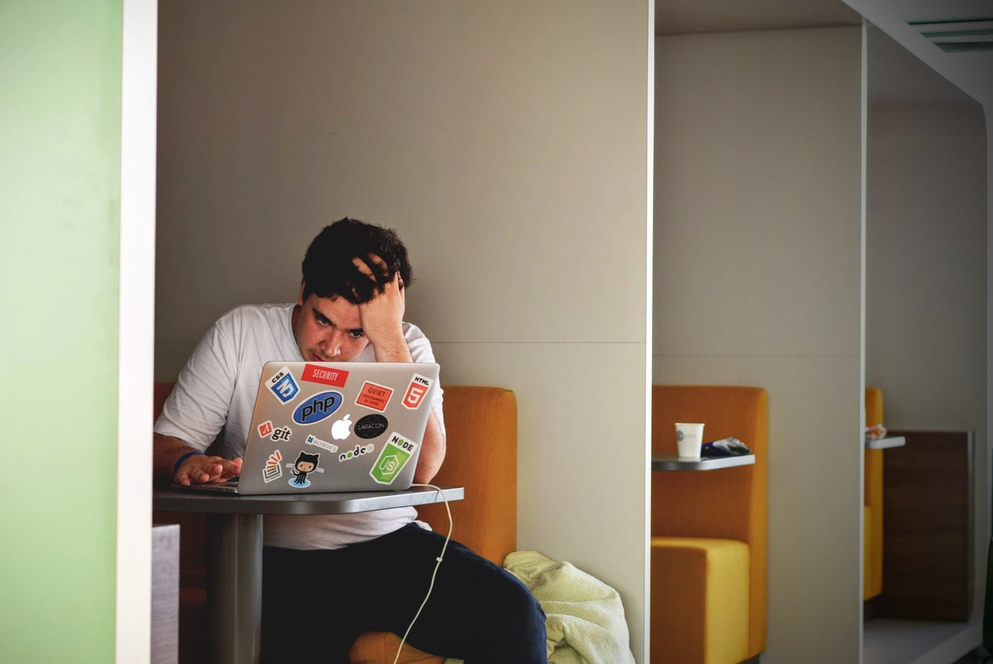 A person sitting in a booth, focused on a laptop covered with various tech and social media stickers, in a modern workspace. They rest their head on their hand, suggesting deep concentration or frustration. A coffee cup is on the table.
