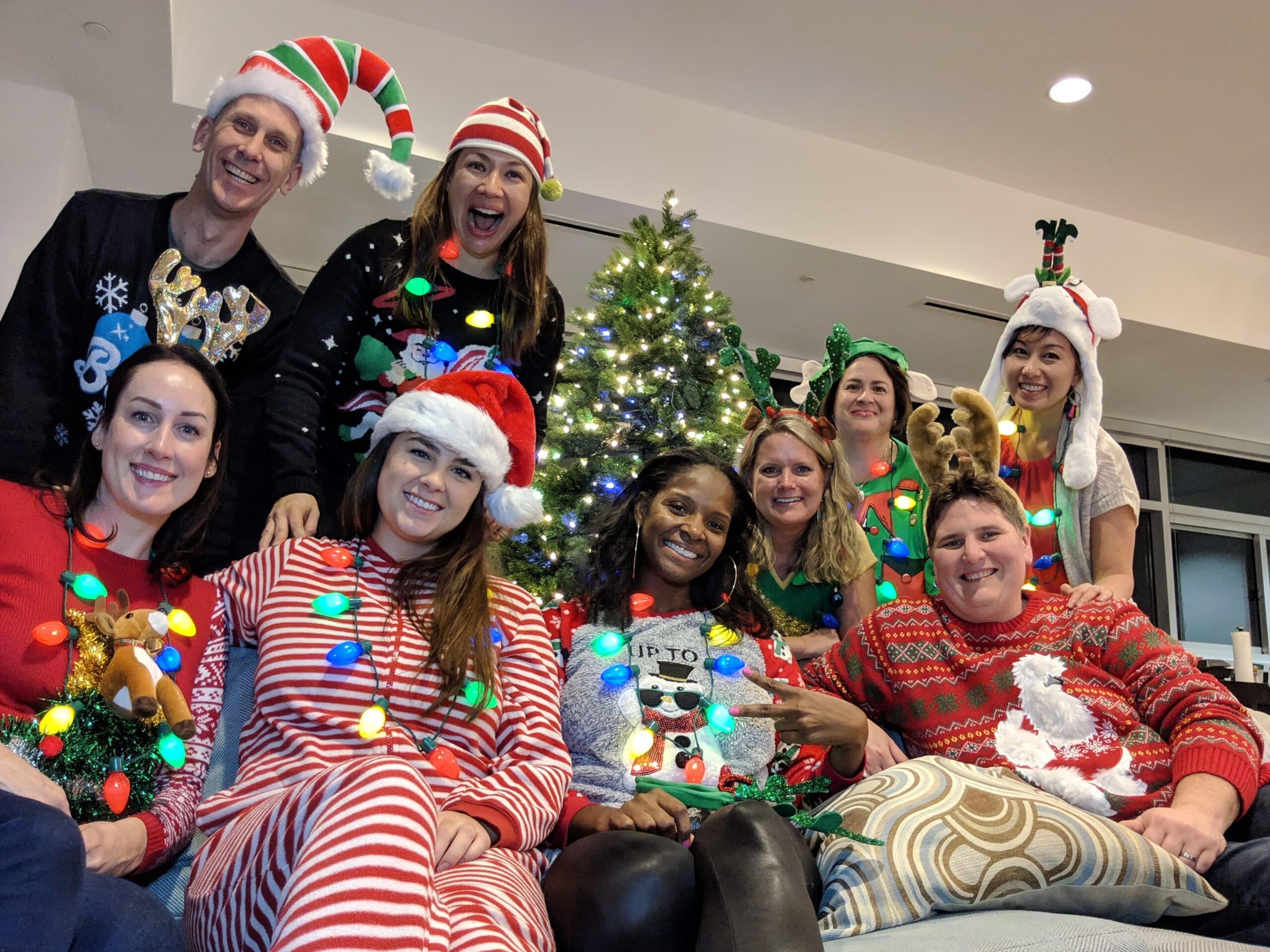 A group of smiling people dressed in festive Christmas sweaters, hats, and pajamas pose together in front of a decorated Christmas tree. They are wearing various holiday-themed accessories, and the atmosphere is joyful and celebratory.